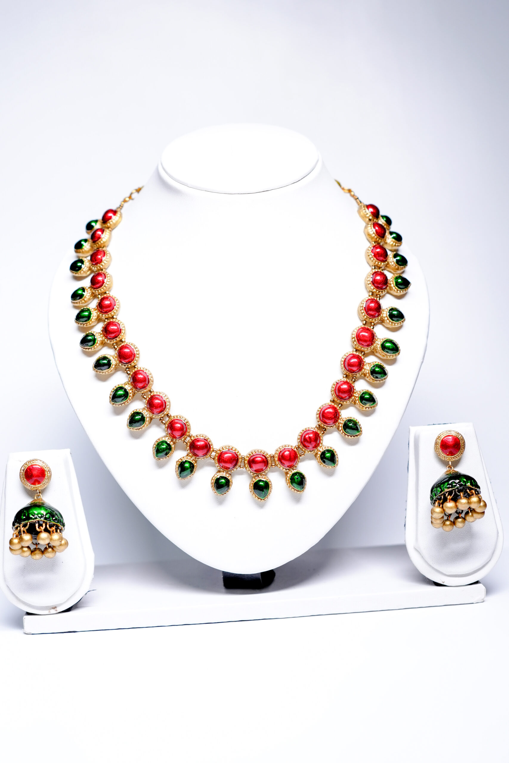 Gold plated necklace with alternating red and green kemp stones and matching jhumka earrings displayed on a white jewellery bust.