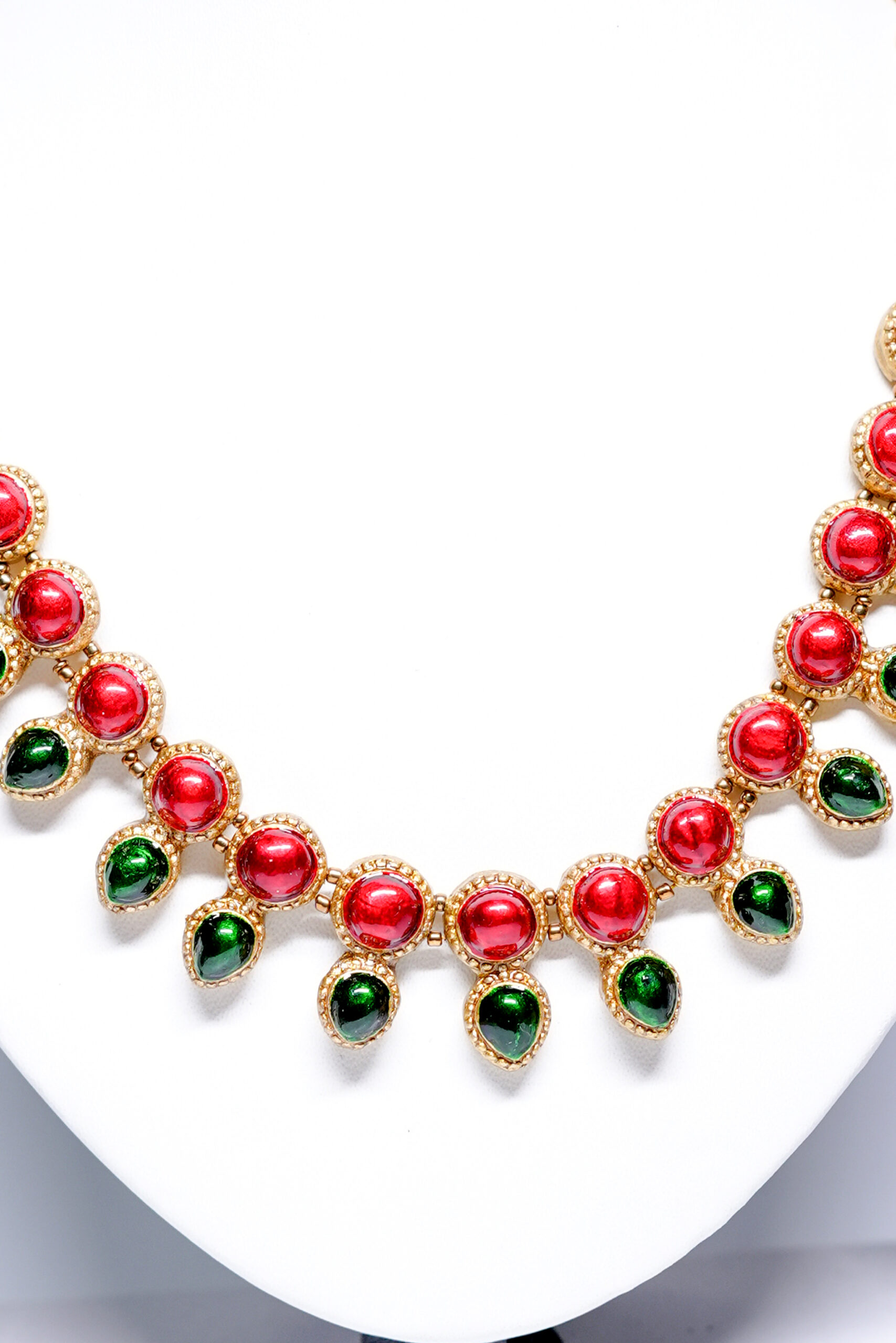 Gold plated necklace with alternating red and green kemp stones displayed on a white jewellery bust, with matching jhumka earrings on either side.