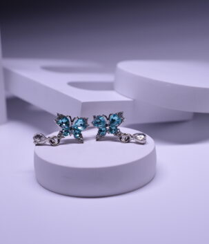 Silver plated butterfly stud earrings with sparkling blue topaz crystals and a hanging clear teardrop, displayed on a white pedestal.