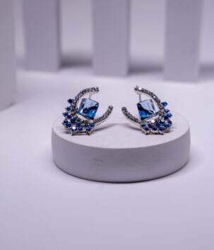 Silver plated crescent moon and star earrings with a bold blue crystal centerpiece and sparkling stone edging, displayed on a white pedestal