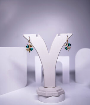 Gold plated four-leaf clover drop earrings featuring emerald green heart crystals and sparkling accents, displayed on a jewellery stand.