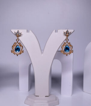 Gold plated blue topaz drop earrings featuring an ornate filigree frame and sparkling crystal accents, displayed on a jewellery stand.