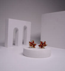 Gold plated maple leaf stud earrings with amber and red crystal detailing, displayed on a white pedestal.