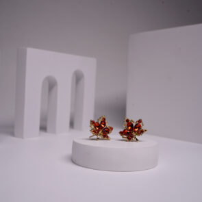 Gold plated maple leaf stud earrings with amber and red crystal detailing, displayed on a white pedestal.