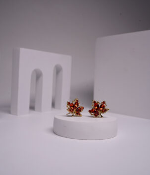 Gold plated maple leaf stud earrings with amber and red crystal detailing, displayed on a white pedestal.