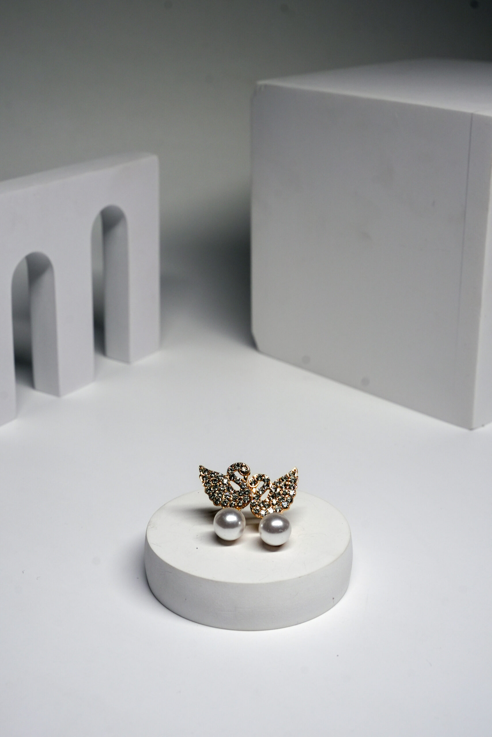 Gold plated swan stud earrings with crystal detailing and a pearl drop, displayed on a white pedestal.