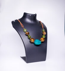 A handcrafted statement necklace with a large turquoise centrepiece, chunky tiger's eye and green jade beads, gold-tone accents, and a brown suede cord, displayed on a black jewellery bust.