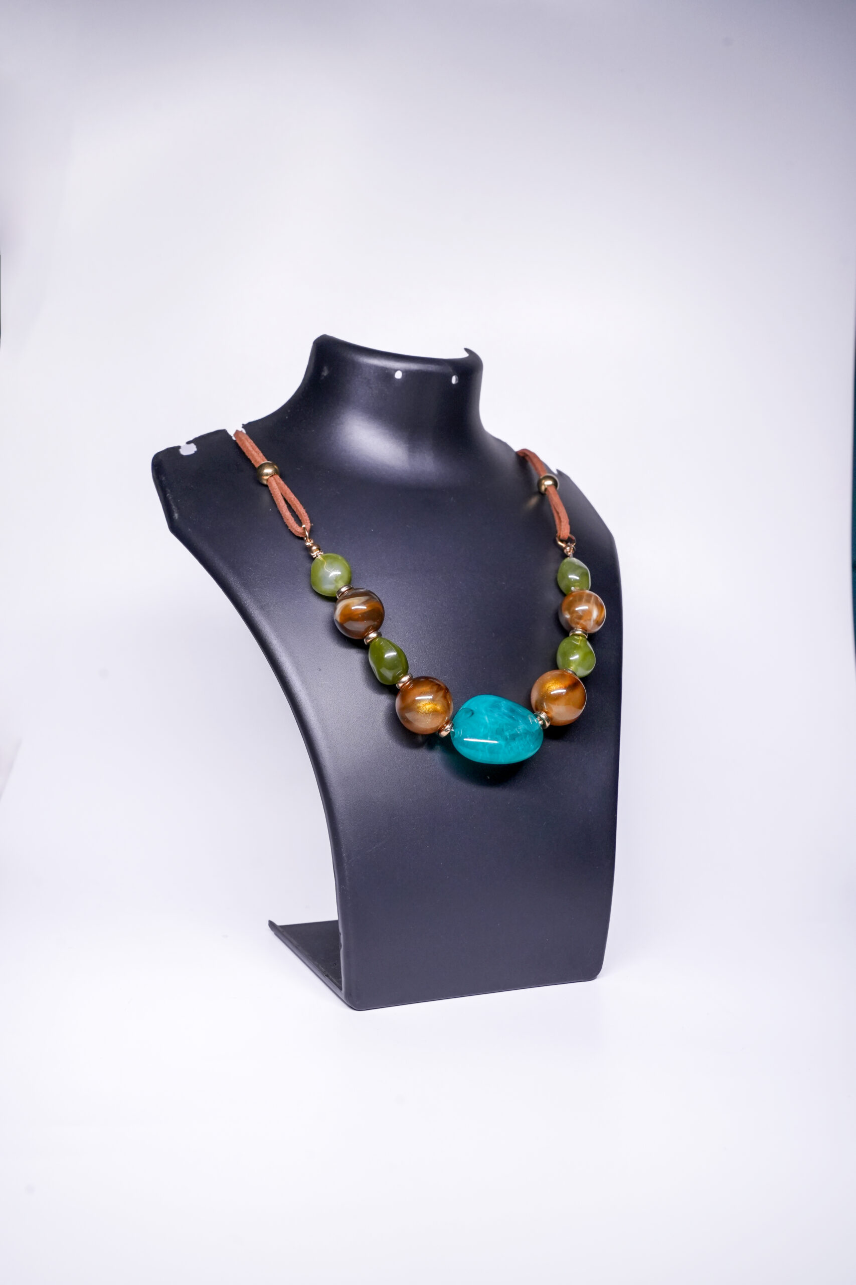 A handcrafted statement necklace with a large turquoise centrepiece, chunky tiger's eye and green jade beads, gold-tone accents, and a brown suede cord, displayed on a black jewellery bust.