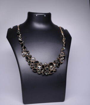 A gold plated statement necklace featuring a dense cluster of black and clear crystal stones, displayed on a black jewellery bust against a grey backdrop