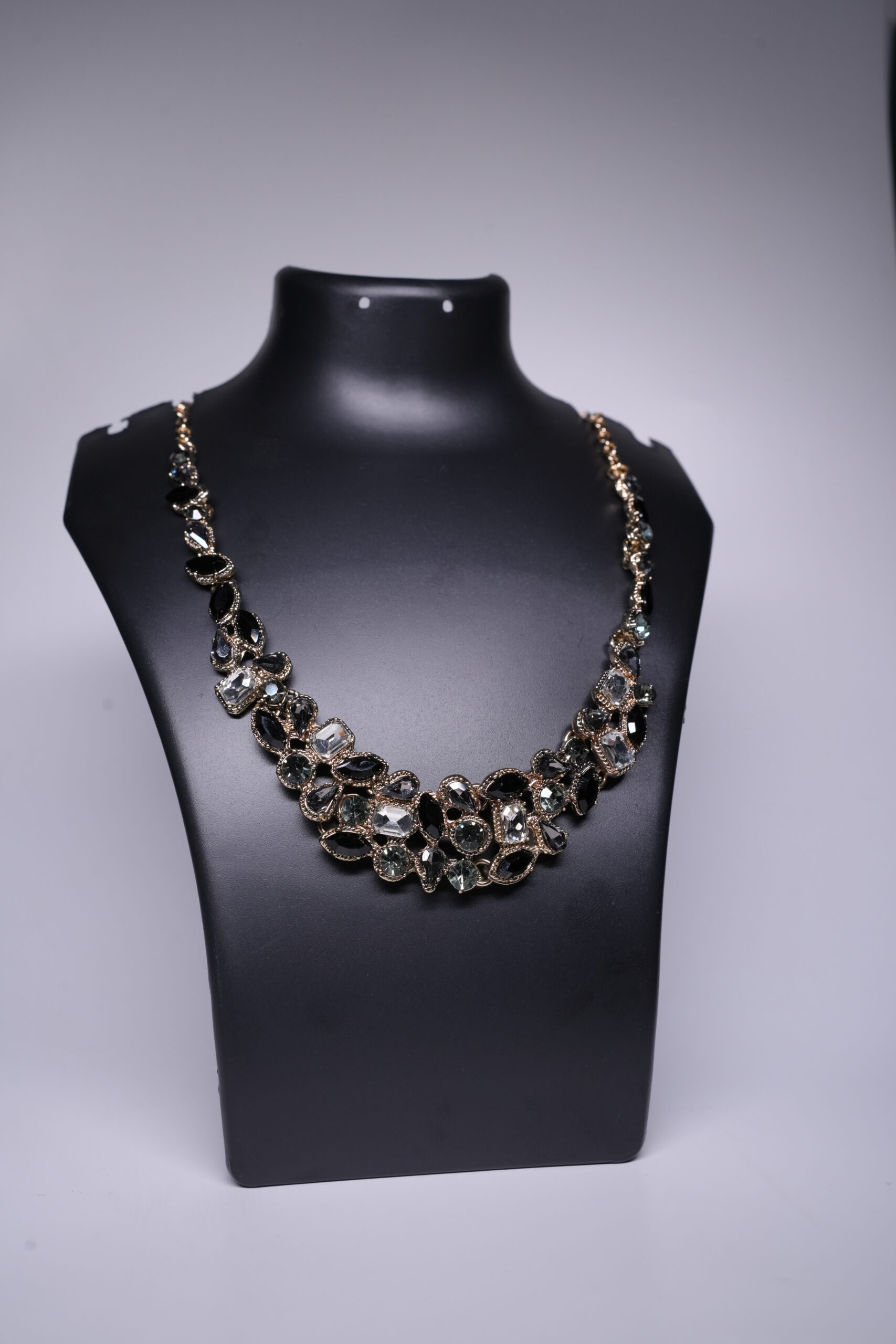 A gold plated statement necklace featuring a dense cluster of black and clear crystal stones, displayed on a black jewellery bust against a grey backdrop