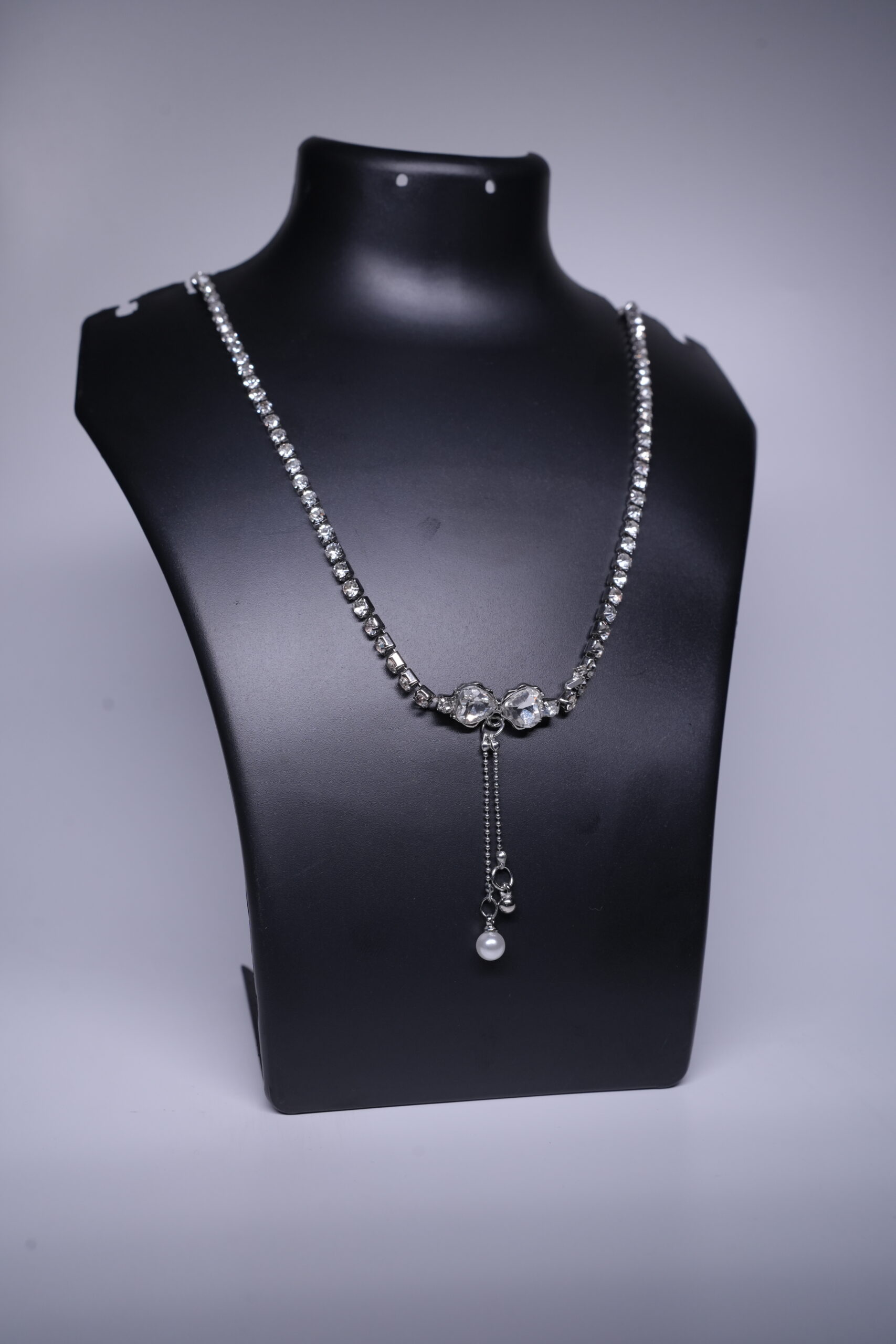 A silver toned CZ tennis necklace with a dainty pearl drop, displayed on a black jewellery bust.