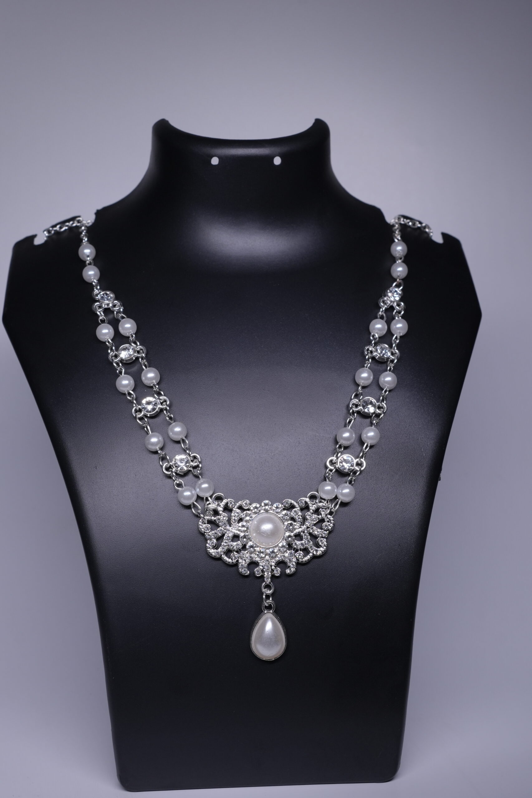 A silver toned bridal necklace with double pearl strands, sparkling CZ stones, and a filigree pendant with a pearl drop, displayed on a black jewellery bust.