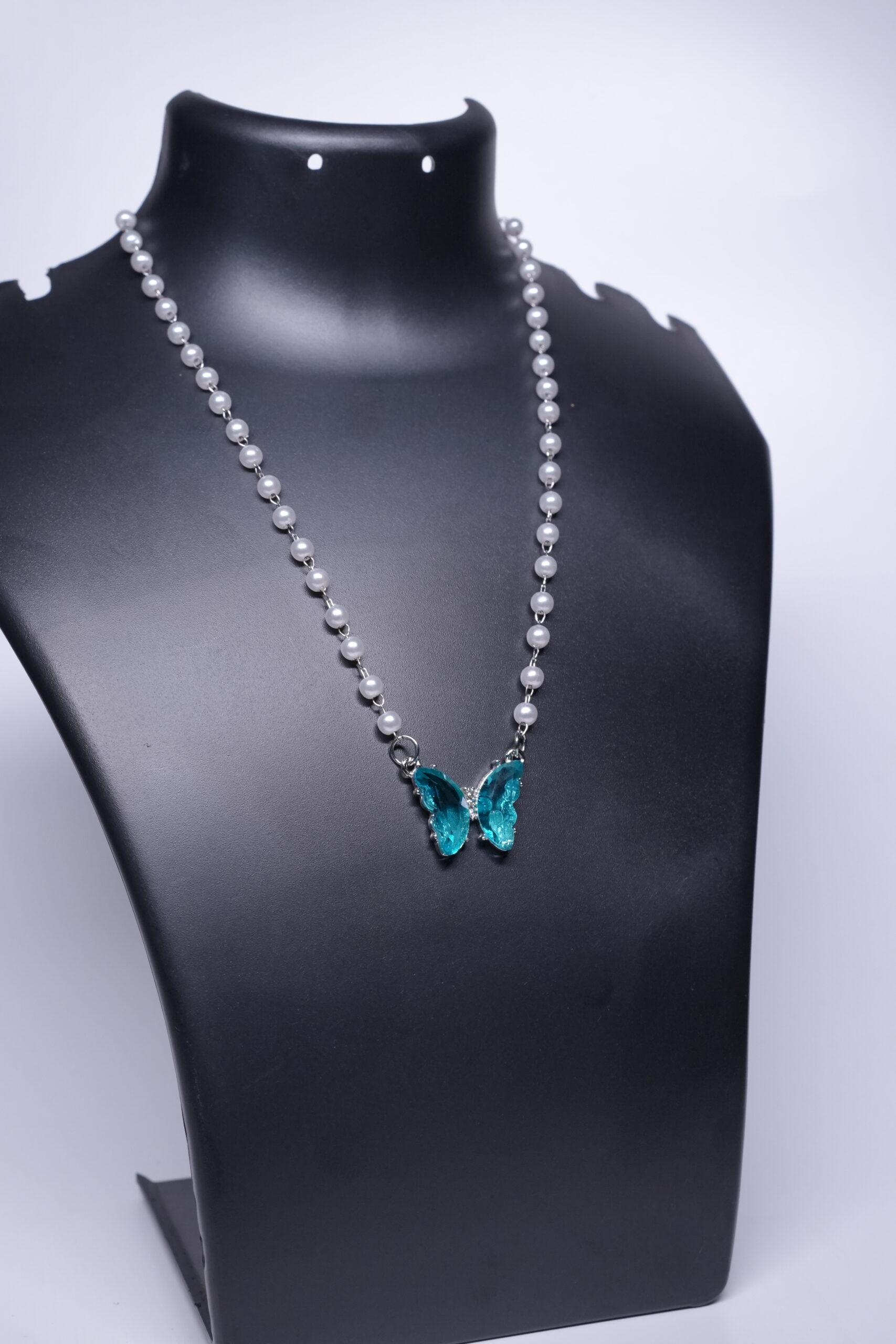 A silver plated faux pearl chain necklace with a sparkling teal crystal butterfly pendant, displayed on a black jewellery bust.