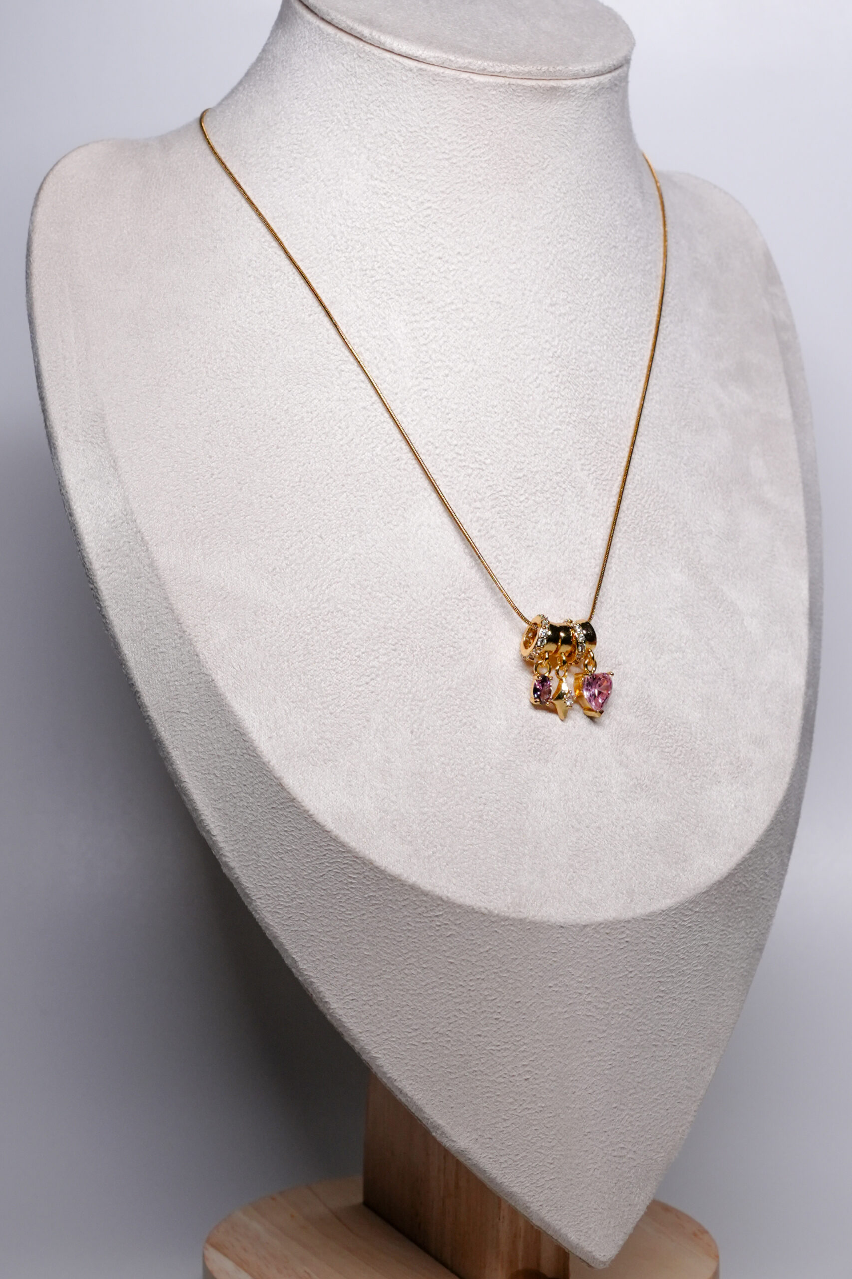 Gold plated snake chain necklace from d2dazzle with pink and lilac zircon heart charms and a gold star charm on sparkling rings, shown on a grey bust stand.
